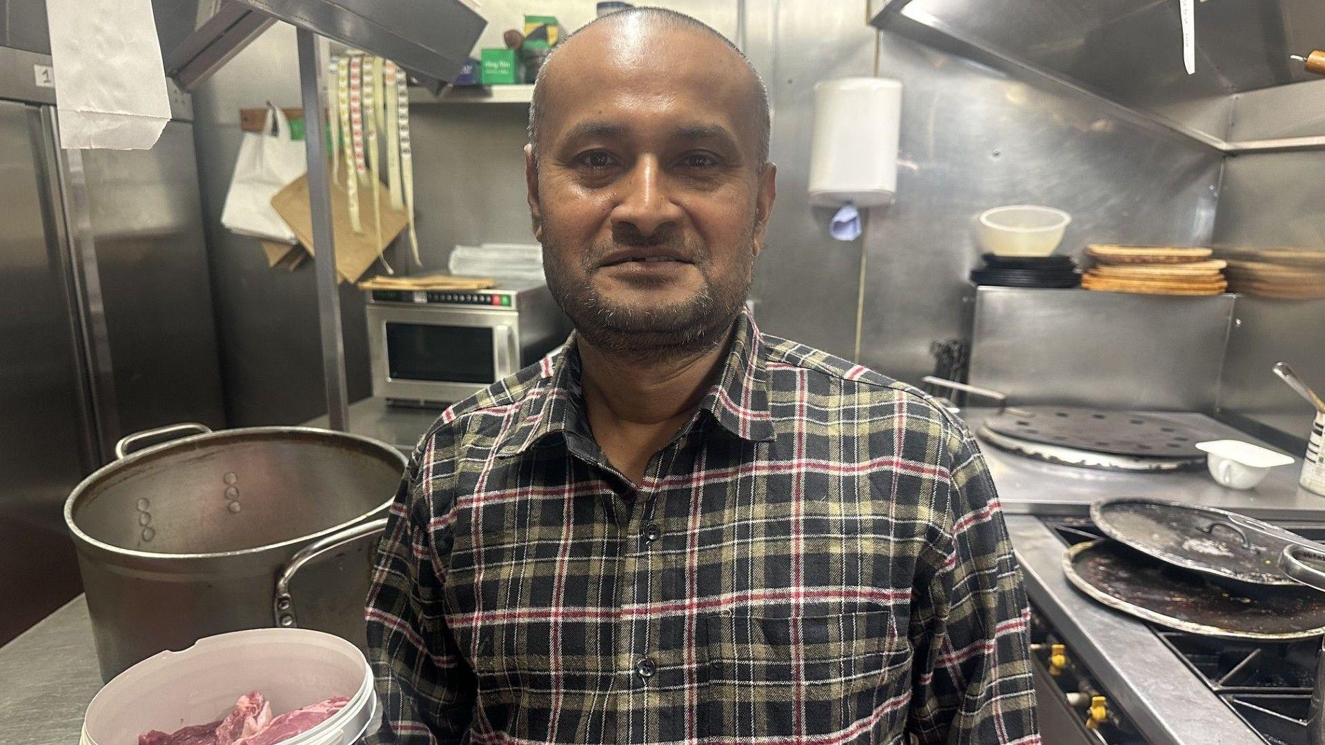 A person wearing a plaid shirt stands in a commercial kitchen next to a container of raw meat. Large cooking pots, a stove, and stainless steel surfaces are visible in the background.